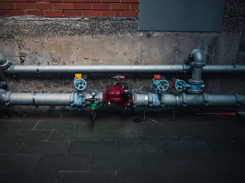 Exposed metal water pipes and control valves running along an exterior brick and concrete wall.