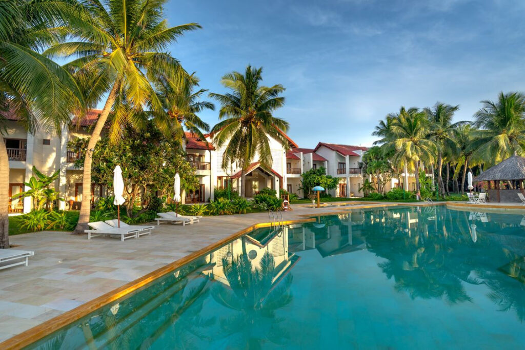 Resort swimming pool surrounded by palm trees, lounge chairs, and white buildings.